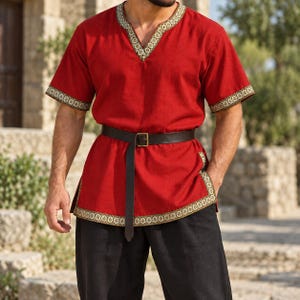 Peut inclure: Une tunique rouge à manches courtes avec une encolure en V et des bords ornés d'un motif circulaire doré et noir. Le vêtement est resserré à la taille par une ceinture en cuir marron foncé dotée d'une boucle métallique, portée avec un pantalon noir.
