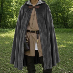 May include: A floor-length dark gray velvet hooded cloak featuring a brown leather clasp at the neck. Underneath, a tan tunic is cinched with a brown leather belt that has a metal buckle and a small brown leather pouch hanging from a cord.