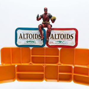 May include: A Deadpool action figure sits atop two Altoids mint tins, one blue and one red, with orange storage containers below. The Altoids tins read "Wintergreen" and "Peppermint".