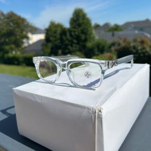 Puede incluir: Gafas transparentes con montura rectangular y detalles decorativos en las patillas. Las lentes tienen un pequeño emblema circular. Las gafas están sobre una caja blanca, con un fondo de árboles y cielo azul.
