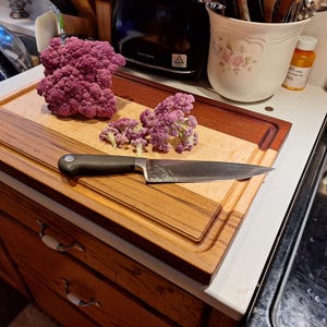 May include: A rectangular wooden cutting board featuring a light-colored center panel framed by dark wood borders with a juice groove. A stainless steel chef knife with a black handle rests on the board alongside several florets of purple cauliflower.