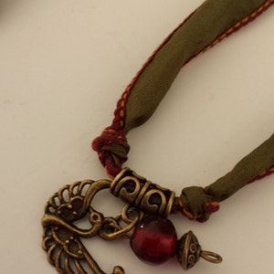 May include: A green fabric cord necklace with a red glass bead and a gold metal bird charm.