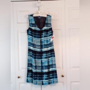 May include: Sleeveless dress with a blue, black, and white plaid pattern. The dress is hanging on a white hanger against a white door. The dress has a V-neck and a tag attached.
