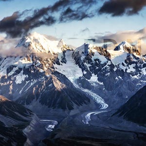 Puede incluir: Fotografía de un paisaje de una cordillera con picos nevados. La imagen captura un valle con un río sinuoso, rodeado de montañas oscuras y rocosas. El cielo es una mezcla de azul y nubes.