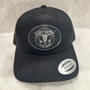May include: Black snapback hat with a mesh back and a curved bill. The front features an oval patch with the text "Casey Wayne & the Rodeo" and a bull skull graphic. A silver sticker is on the side.