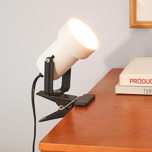 May include: A white and black clip-on spotlight lamp with a bright light. The lamp is attached to a wooden surface. A black power cord extends from the lamp. The lamp is angled towards the viewer.