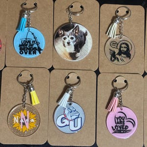 Puede incluir: Una colección de llaveros con varios diseños. Los llaveros incluyen un dinosaurio morado, un perro, un girasol y una rana. Algunos llaveros tienen texto, como "Love it when she bends over" y "You are loved."