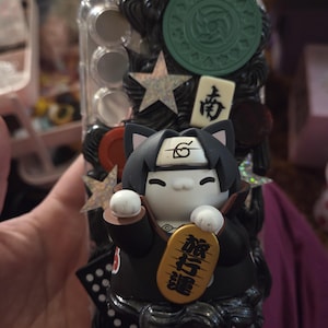 May include: A clear phone case decorated with a black cat figurine wearing a black cloak and headband. The cat holds a gold-colored sign with Japanese text. The case also features star-shaped embellishments and a green circular design.