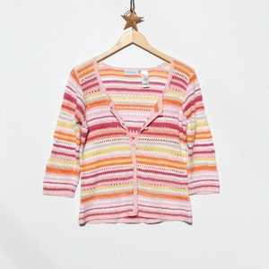 Pu&ograve; includere: Un cardigan a righe rosa, arancioni e gialle con maniche a tre quarti. Il maglione &egrave; abbottonato e appeso a un'appendiabiti di legno su sfondo bianco. Il nome del marchio "villager" &egrave; visibile sull'etichetta.
