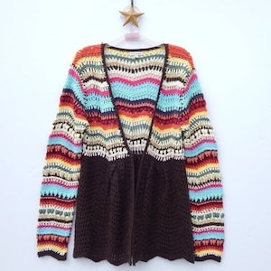 Può includere: Un cardigan in crochet marrone con strisce colorate in tonalità di rosso, arancione, giallo, verde, blu e rosa. Il cardigan ha un collo a V e una chiusura anteriore.