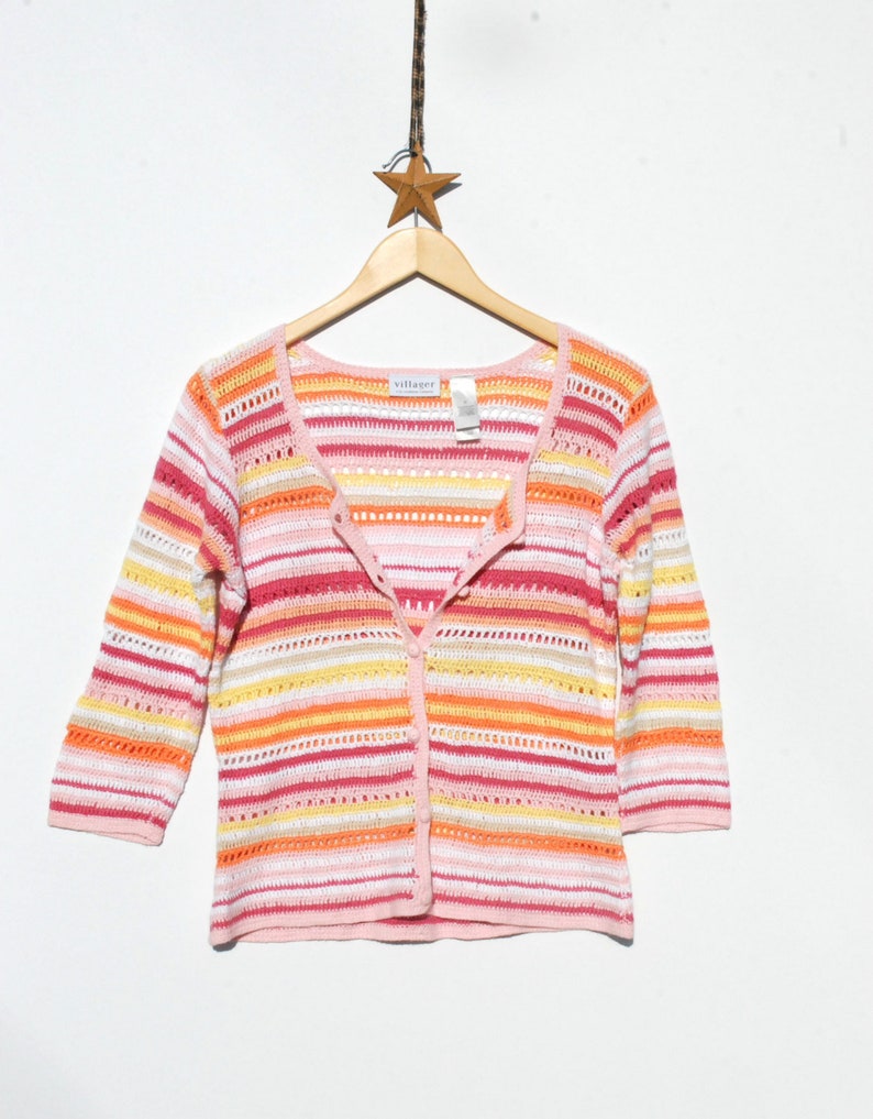 Pu&ograve; includere: Un cardigan a righe multicolore con maniche a tre quarti. Il maglione presenta righe orizzontali nelle tonalit&agrave; del rosa, dell'arancione, del giallo e del bianco. Ha una chiusura a bottoni sul davanti ed &egrave; appeso a una gruccia di legno.