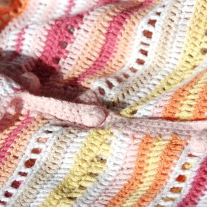 Pu&ograve; includere: Primo piano di una coperta all'uncinetto fatta a mano in una variet&agrave; di colori. La coperta presenta un motivo a righe con sfumature di rosa, bianco, giallo e arancione. Il lavoro all'uncinetto ha un aspetto strutturato con piccoli fori.