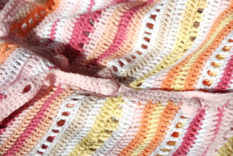 Pu&ograve; includere: Primo piano di una coperta all'uncinetto fatta a mano in una variet&agrave; di colori. La coperta presenta un motivo a righe con sfumature di rosa, bianco, giallo e arancione. Il lavoro all'uncinetto ha un aspetto strutturato con piccoli fori.