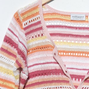 Pu&ograve; includere: Un cardigan all'uncinetto a righe dai colori pastello con scollo a V e chiusura con bottoni. La tavolozza dei colori comprende sfumature di rosa, bianco, giallo e arancione. L'etichetta recita "villager a liz claiborne company". Il cardigan &egrave; appeso a un'appendiabiti di legno.