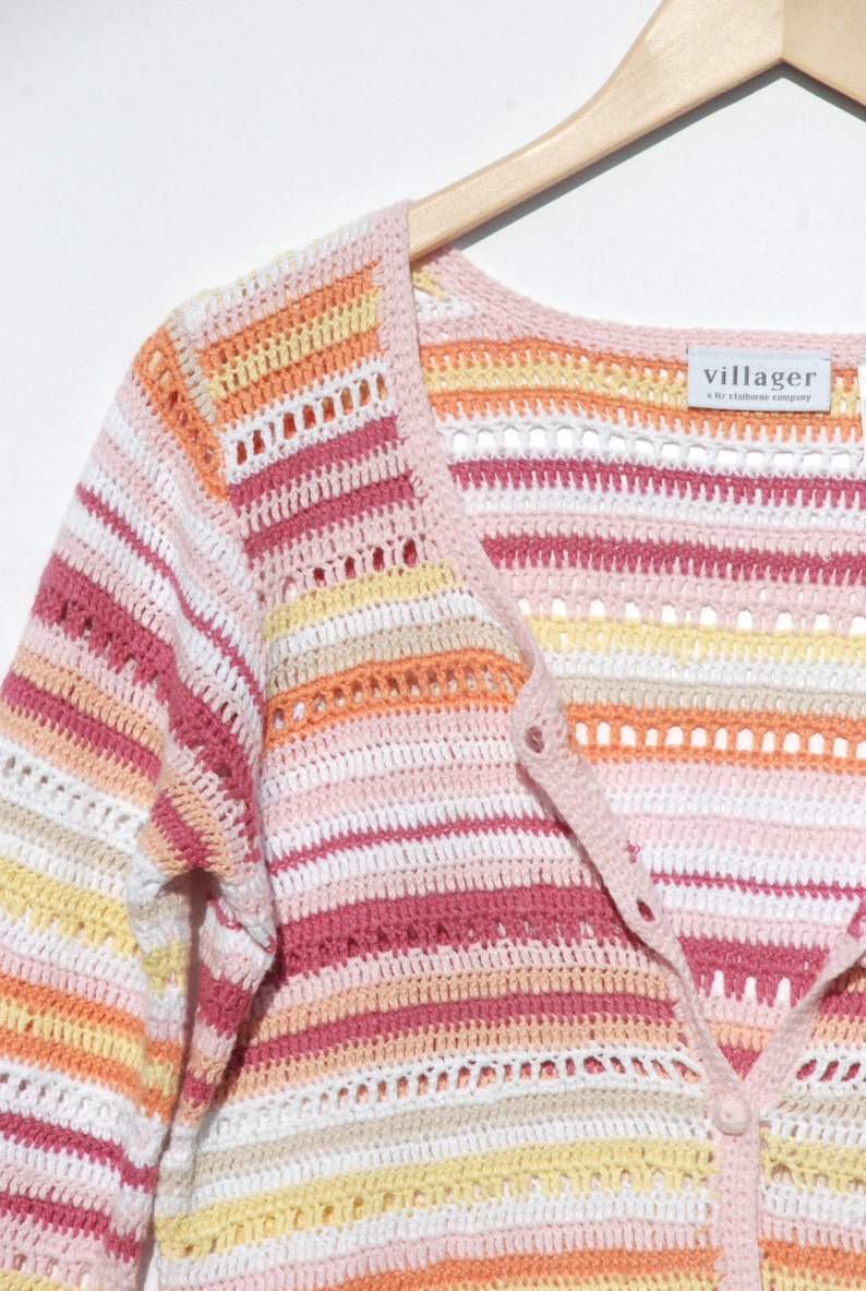 Pu&ograve; includere: Un cardigan all'uncinetto a righe dai colori pastello con scollo a V e chiusura con bottoni. La tavolozza dei colori comprende sfumature di rosa, bianco, giallo e arancione. L'etichetta recita "villager a liz claiborne company". Il cardigan &egrave; appeso a un'appendiabiti di legno.
