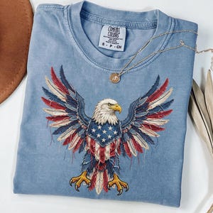 May include: A light blue crew neck t-shirt featuring a graphic of a bald eagle with wings spread wide. The eagle's feathers are stylized with red, white, and blue patterns resembling the American flag. The shirt has a Comfort Colors brand label.