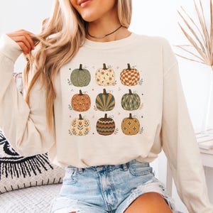May include: A cream-colored long-sleeve sweatshirt featuring a graphic print of nine pumpkins arranged in a grid. Each pumpkin displays a unique pattern, including wavy lines, celestial motifs, checkered squares, floral designs, and starburst shapes.