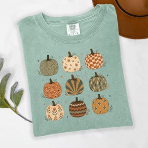 May include: A sage green t-shirt featuring a graphic print of nine pumpkins, each with a unique pattern including checkers, suns, moons, and floral motifs. The shirt has a Comfort Colors label at the neck and a crew neckline with short sleeves.