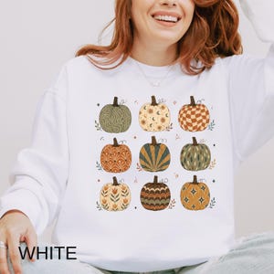 May include: A white long-sleeve sweatshirt featuring a graphic print of nine pumpkins arranged in a grid. Each pumpkin displays a unique pattern, including wavy lines, celestial motifs, checkered squares, floral designs, and sunburst rays in earthy tones.