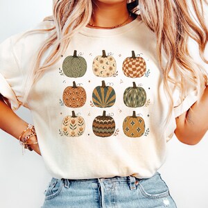 May include: A cream-colored t-shirt featuring a graphic print of nine pumpkins arranged in a grid. Each pumpkin displays a unique pattern, including wavy lines, celestial motifs, checkered squares, floral designs, and starbursts in earthy tones.