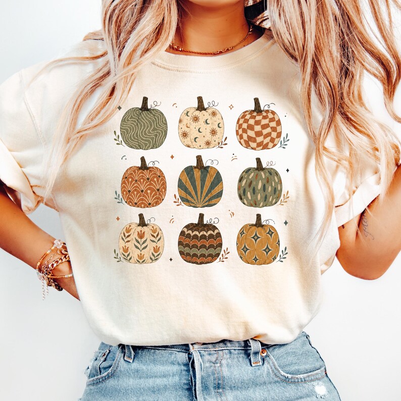 May include: A cream-colored t-shirt featuring a graphic print of nine pumpkins arranged in a grid. Each pumpkin displays a unique pattern, including wavy lines, celestial motifs, checkered squares, floral designs, and starbursts in earthy tones.
