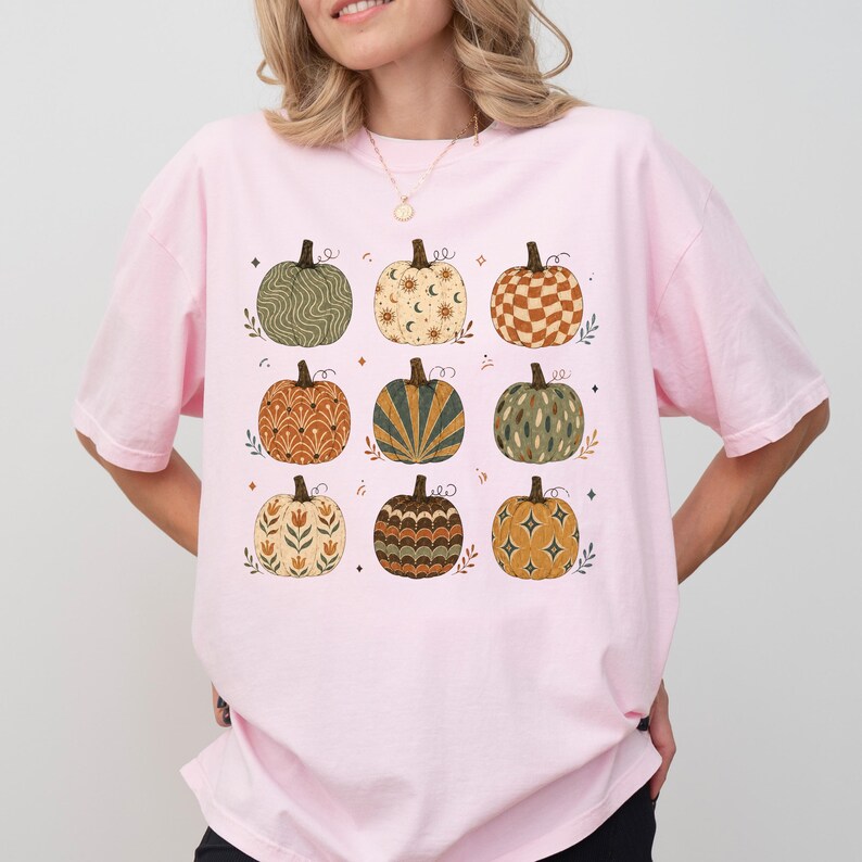 May include: A light pink short-sleeve t-shirt featuring a graphic print of nine pumpkins arranged in a grid. Each pumpkin displays a unique pattern, including wavy lines, celestial motifs, checkered squares, floral designs, and geometric starbursts.