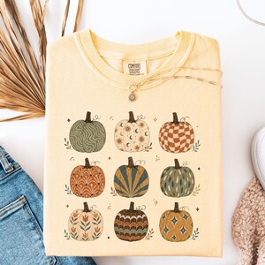 May include: A pale yellow short-sleeve t-shirt featuring a graphic print of nine pumpkins arranged in a grid. Each pumpkin displays a unique pattern, including wavy lines, celestial motifs, checkered squares, floral designs, and geometric shapes.