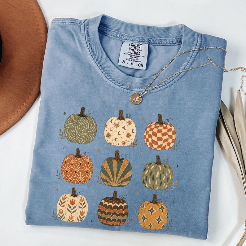 May include: A blue short-sleeved t-shirt featuring a graphic print of nine pumpkins, each with a unique pattern including waves, celestial symbols, checkers, sunbursts, and floral motifs. The shirt has a Comfort Colors brand label at the neck.