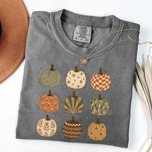 May include: A charcoal gray short-sleeve t-shirt featuring a graphic print of nine pumpkins in various patterns including waves, suns, moons, checkers, and floral motifs. The shirt has a crew neckline and a Comfort Colors brand label at the inner neck.