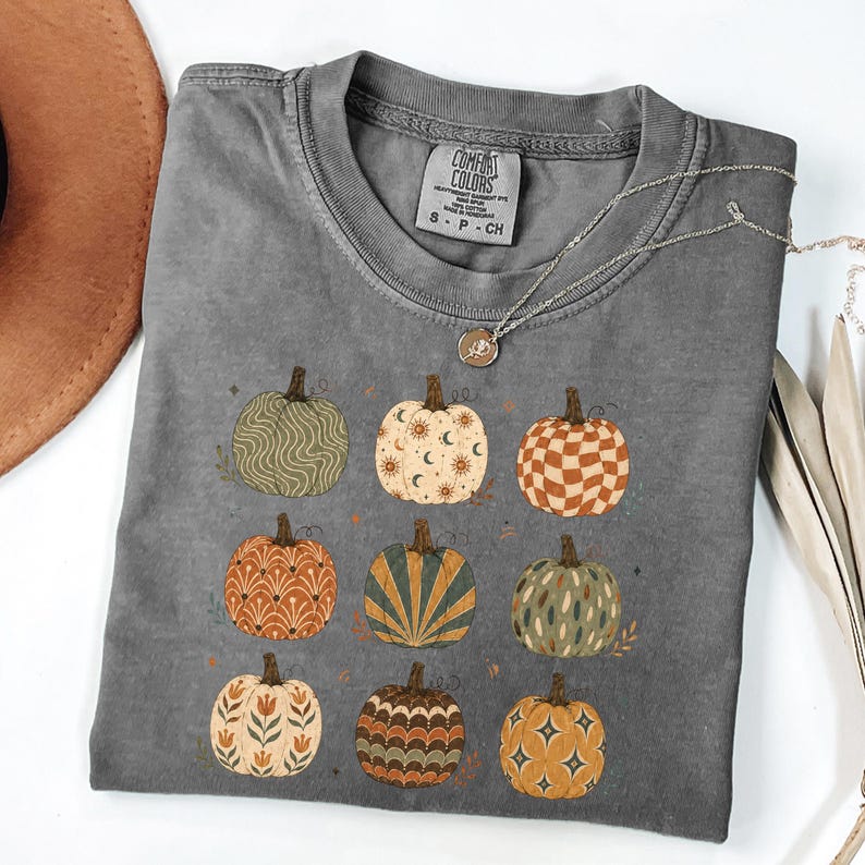May include: A charcoal gray short-sleeve t-shirt featuring a graphic print of nine pumpkins in various patterns including waves, suns, moons, checkers, and floral motifs. The shirt has a crew neckline and a Comfort Colors brand label at the inner neck.