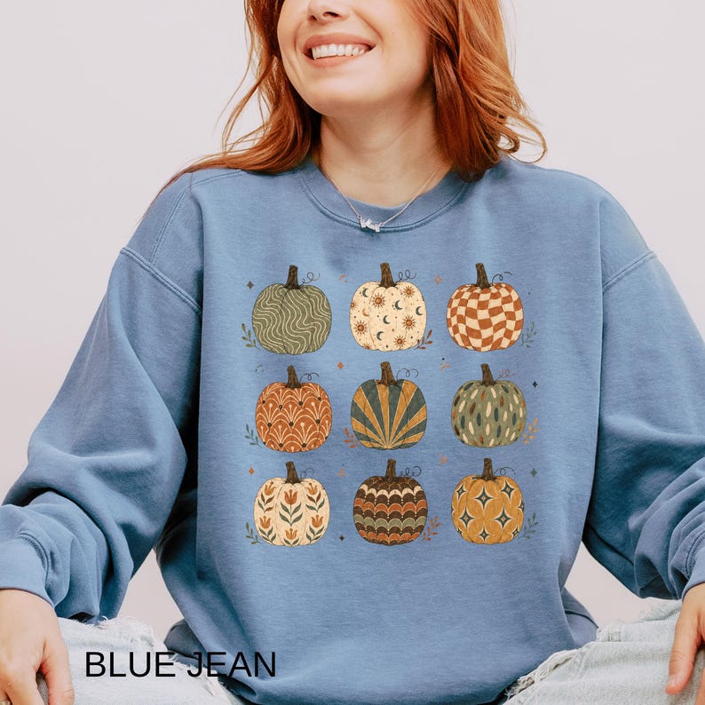 May include: A dusty blue crewneck sweatshirt featuring a graphic print of nine pumpkins arranged in a three-by-three grid. Each pumpkin displays a unique pattern, including wavy lines, celestial motifs, checkered squares, and floral designs.