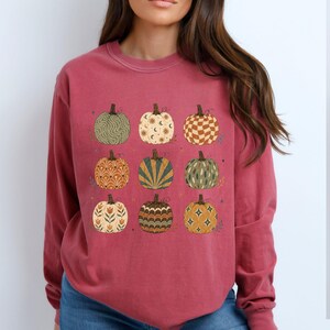 May include: A long-sleeve crewneck sweatshirt in a muted red color featuring a graphic print of nine pumpkins arranged in a grid. Each pumpkin displays a unique pattern, including wavy lines, celestial motifs, checkered squares, and floral designs.
