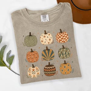 May include: A taupe short-sleeve t-shirt with a crew neckline and a Comfort Colors brand label. The front features a graphic print of nine pumpkins in various patterns, including wavy lines, celestial moons, checkers, floral motifs, and starbursts.