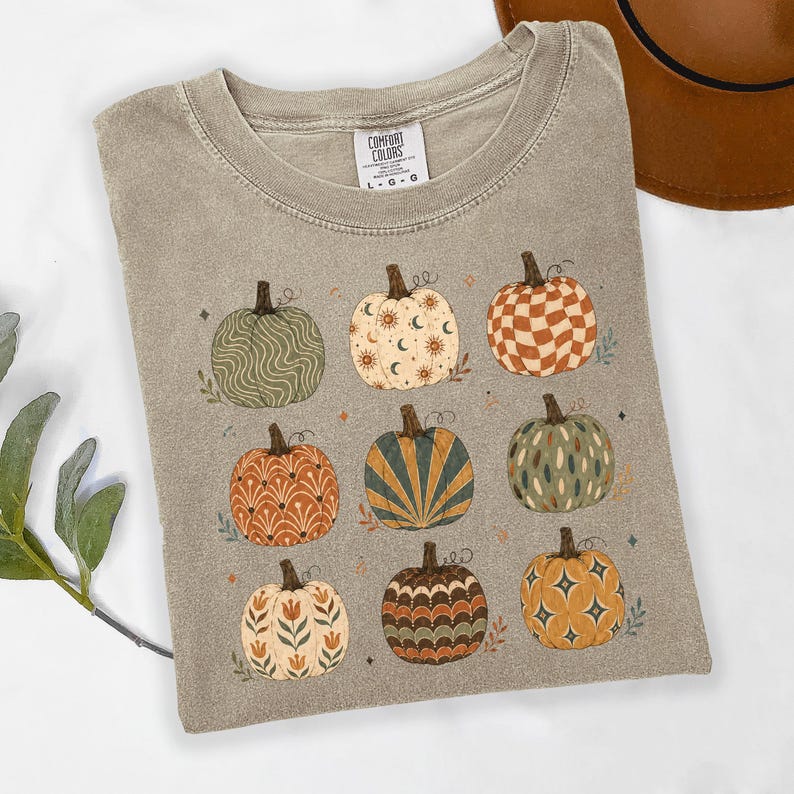 May include: A taupe short-sleeve t-shirt with a crew neckline and a Comfort Colors brand label. The front features a graphic print of nine pumpkins in various patterns, including wavy lines, celestial moons, checkers, floral motifs, and starbursts.