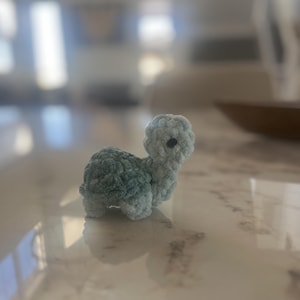 Puede incluir: Una pequeña tortuga de ganchillo hecha a mano en tonos azules y verdes. La tortuga tiene un aspecto texturizado y afelpado con un ojo oscuro. El animal artesanal está sobre una superficie reflectante.