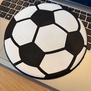 Op de afbeelding: Een ronde muismat in de vorm van een voetbal, zwart en wit. Het heeft het klassieke patroon van vijfhoeken en zeshoeken van een voetbal. De muismat ligt op een laptoptoetsenbord. Gemaakt van zacht materiaal.