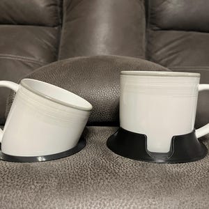 May include: Two white ceramic mugs with grey trim, set in black cup holders. The mugs have a simple design and are placed on a grey leather surface, suggesting a living room setting. The mugs are ready to be filled with tea or coffee.