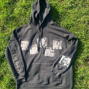 May include: Dark grey hoodie with a front pocket and hood. The hoodie features patches with black and white images of characters and a knife. The hoodie is laying on green grass.