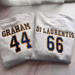 Op de afbeelding: Twee lichtgrijze sweaters met ronde hals en geborduurde letters en cijfers. De linker sweater toont de naam GRAHAM boven het getal 44. De rechter sweater toont de naam DI LAURENTIS boven het getal 66. Alle tekst en cijfers zijn donkerblauw met gouden omlijning.