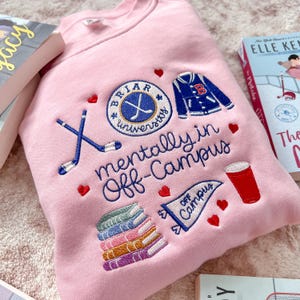 Op de afbeelding: Een lichtroze sweater met ronde hals voorzien van borduursels waaronder een Briar University logo, een varsity jack, gekruiste hockeysticks, een stapel kleurrijke boeken, een rode beker, een wimpel en kleine rode hartjes met de tekst mentally in Off-Campus.