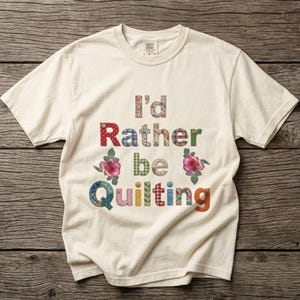 May include: A cream-colored short-sleeve t-shirt featuring the text I'd Rather be Quilting. Each letter is filled with a different colorful fabric pattern, including floral, plaid, and polka dot designs, accented by two pink rose illustrations.