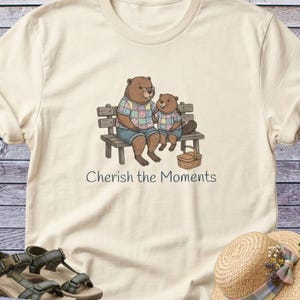 May include: A cream-colored t-shirt featuring a graphic of two brown beavers sitting on a wooden bench. The beavers wear colorful patchwork shirts and blue shorts. A small picnic basket sits on the ground. The text reads, Cherish the Moments.