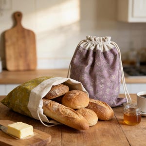 May include: A wooden table displays a variety of baked goods, including baguettes and round loaves, inside a yellow and cream bread bag. A matching purple floral drawstring bag sits nearby. A small jar of honey and a cup of coffee complete the scene.