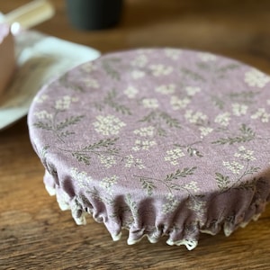 May include: A round, lavender fabric food cover with a floral pattern of white flowers and green leaves. The cover has a ruffled edge with white lace trim. The cover is designed to fit over a bowl or dish.