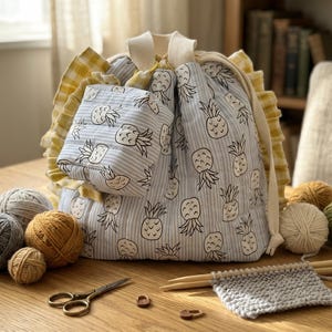 May include: A fabric drawstring project bag featuring a light blue and white striped pattern with black pineapple illustrations. The bag includes yellow and white gingham ruffled trim, a small matching pouch, yarn balls, knitting needles, and scissors.
