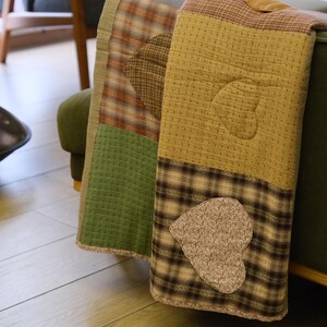 May include: A quilted throw blanket featuring a patchwork design with alternating squares of solid olive green, brown and tan plaid, and textured mustard yellow fabric. The blanket includes stitched heart-shaped appliques in brown and tan.