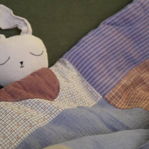 Può includere: Un coniglio di peluche con la testa grigio chiaro e tratti del viso ricamati, che riposa sotto una trapunta patchwork. La trapunta presenta vari quadrati di tessuto nelle tonalità del blu, viola e marrone chiaro, con motivi a righe, quadri e fiori.