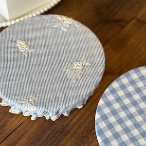 Op de afbeelding: Twee ronde stoffen komhoezen. Eén is blauw en wit gestreept met witte geborduurde bloemenaccenten en een geschulpte kanten rand. De andere is blauw en wit geruit. Beide liggen op een houten oppervlak.