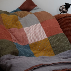 May include: A patchwork quilt featuring a grid of square fabric panels in various colors including mustard yellow, terracotta, sage green, dusty blue, and light pink, with some squares displaying small white dot patterns and gingham checks.