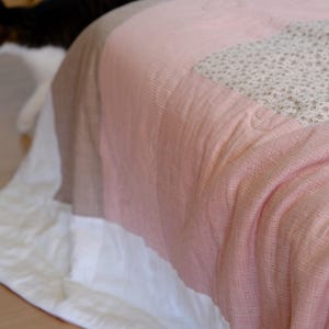 Może przedstawiać: Narzuta typu patchwork składająca się z połączenia gładkich paneli z tkaniny w kolorze różowym, białym i brązowym oraz sekcji w drobny kwiatowy wzór. Materiał ma widoczną tkaną fakturę i jest narzucony na powierzchnię, ukazując liczne zszyte ze sobą elementy.